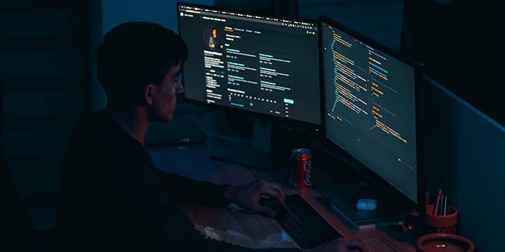 A man sat in a dark room, working on some code on a three-screen setup