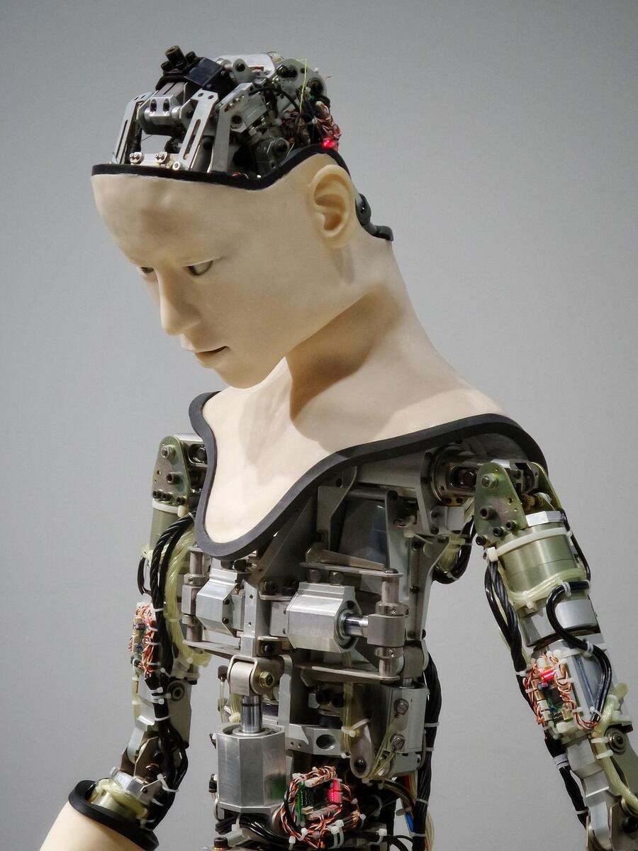A human shaped robot with exposed wires on its body, arms and head but a realistic looking face