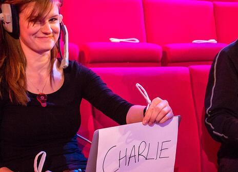 Michelle Reeve holding up a sign that says 'Charlie' next to a cameraman who looks intently at something off camera