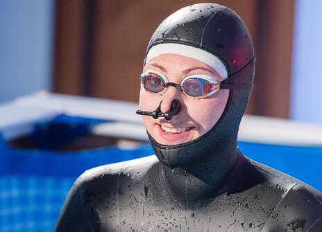 person in full wetsuit with swim goggles and a nose clip sitting in a paddling pool
