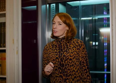 Hannah Fry walking out of a lift, with bookshelves either side of her