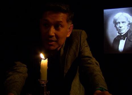 Saiful Islam leaning in front of a lit candle, with a portrait of Michael Faraday in the background