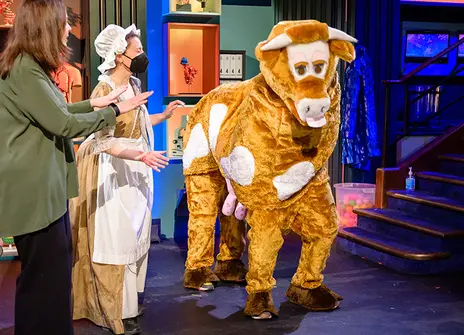 a pantomime cow is at the Royal Institution lecture theatre, next to her are a woman in a milkmaid outfit and another person in a green shirt