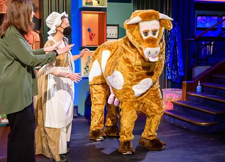 a pantomime cow is at the Royal Institution lecture theatre, next to her are a woman in a milkmaid outfit and another person in a green shirt