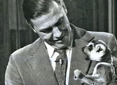 David Attenborough looking down at a lemur that he is holding  