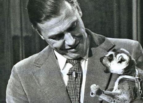 David Attenborough looking down at a lemur that he is holding  