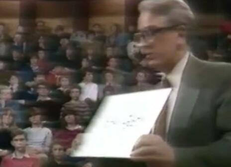 David Phillips holding a sheet of paper in the Ri theatre, a crowd of children watching from their seats