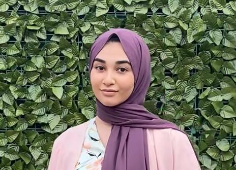 A teenage girl wearing a mauve headscarf, white floral dress and pink top, standing in front of a green leafy background, 