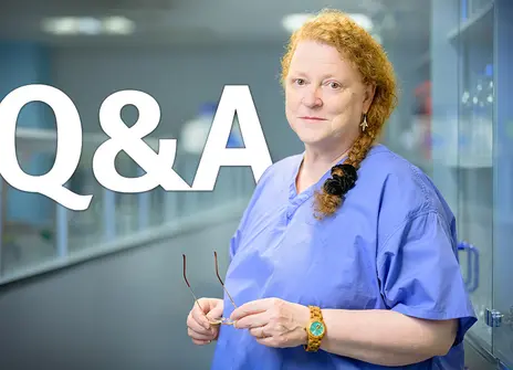 Sue Black standing in a lab, wearing blue scrubs and holding her glasses
