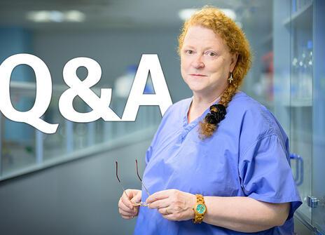 Sue Black standing in a lab, wearing blue scrubs and holding her glasses