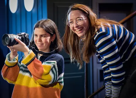 Mia is wearing an orange striped jumper, holding a camera. Behind her, a woman with long brown hair laughs.