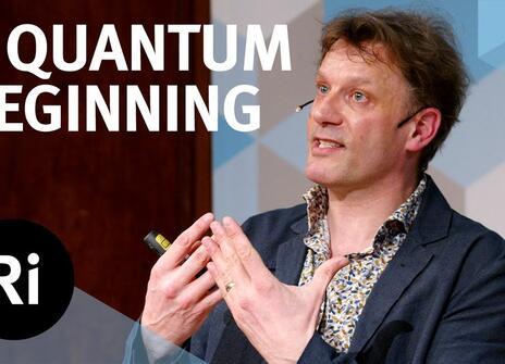 A presenter stands on a stage with his hands slightly raised. The words 'A QUANTUM BEGINNING' are written above, along with the Royal Institution Logo.
