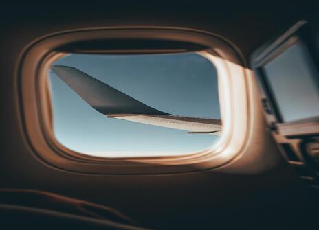 An airplane wing viewed through an airplane window mid-flight, with blue sky in the background.