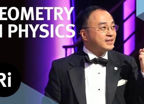 A presenter in black tie stands on a stage. The words 'GEOMETRY IN PHYSICS' are written above, along with the Royal Institution Logo.
