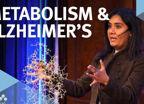 The speaker faces the front of the stage, hands slightly raised above a model of a chemical structure. Overlayed are the words 'METABOLISM & ALZHEIMER'S' alongside the Royal Institution's logo.