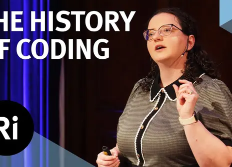 The speaker faces the front of the stage, hands slightly raised. Overlayed are the words 'THE HISTORY OF CODING' alongside the Royal Institution's logo.