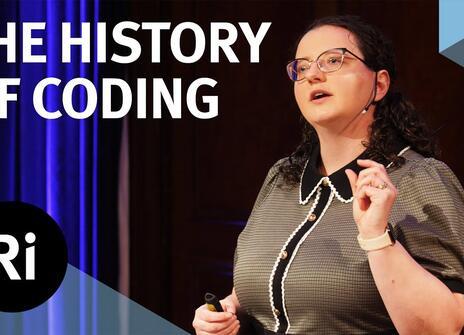The speaker faces the front of the stage, hands slightly raised. Overlayed are the words 'THE HISTORY OF CODING' alongside the Royal Institution's logo.