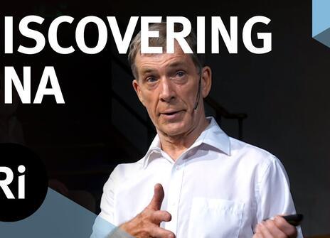 A presenter stands on a stage with his hands slightly raised. The words 'DISCOVERING DNA' are written above, along with the Royal Institution Logo.