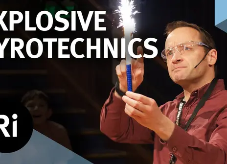 A presenter on stage holds a stick which has sparks shooting out from one end. The words 'EXPLOSIVE PYROTECHNICS' are written above, along with the Royal Institution Logo.