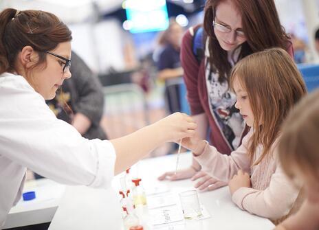 Young people work together with a female scientist dressed in a lab coat, looking at science equipment