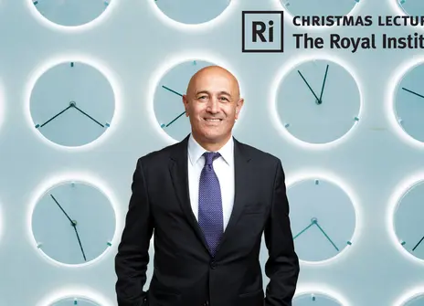 Jim Al-Khalili in front of a teal wall of analogue clocks