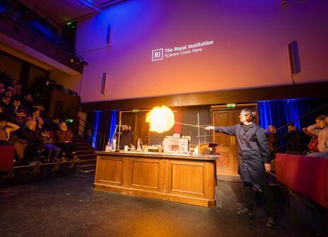 a person is holding out a long stick with a flame on the end. it is touching a balloon which has turned into a yellow ball of fire. 