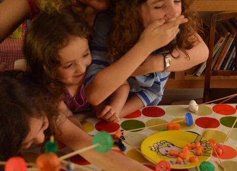three young girls at a kitchen table building a sculpture out of wooden skewers, pieces of carrot and marshmallows