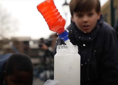 a small fruit juice bottle is being propelled out of a glass jar by a white jet of liquid as a young boy looks on