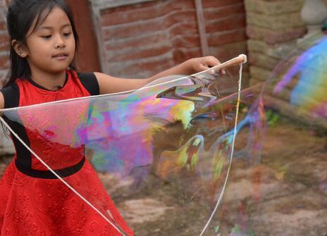 young girl in red dress holding two wooden spoons and some string tied in a loose circle around them from which emerges a giant bubble