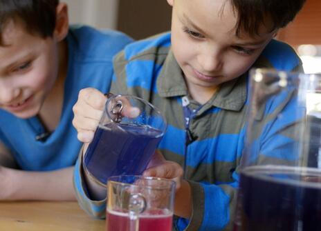 one young boy is pouring a purple liquid into a pink liquid while another boy looks on