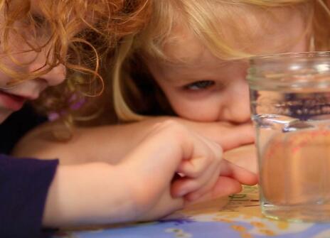 two young girls leaning over a table looking at an unpeeled egg floating in a glass jar