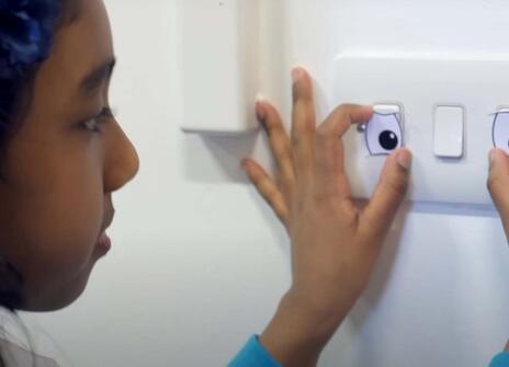 a girl is holding up two cardboard eyes over a light switch making it looks as if it's a face