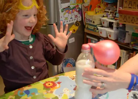 a young girl with curly red hair has her hands up in delighted surprise as she looks at a balloon self-inflating