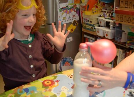 a young girl with curly red hair has her hands up in delighted surprise as she looks at a balloon self-inflating