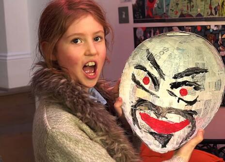 a girl holds up a large papier maché mask