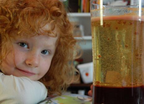 red curly haired young girl smiles to the camera, next to her is a glass filled with oil and some red liquid