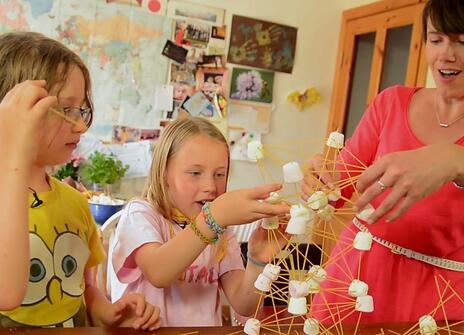 two small children and a woman in a red dress are building a tower out of spaghetti and marshmallows