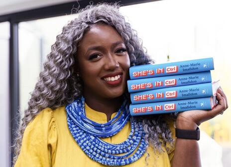 A woman is holding copies of the book 'She's in CTRL' up to her face. She is smiling and wearing a yellow top. 