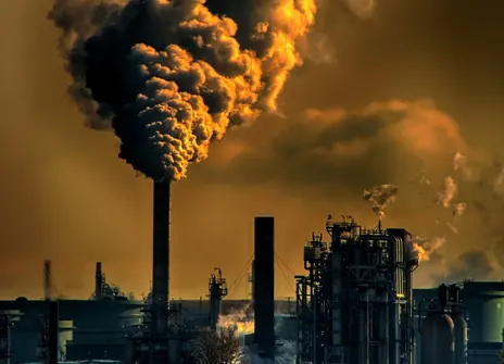 There is a landscape of chimney towers with lots of smoke coming out of them. The picture is a brown and yellow colour to show air pollution. 