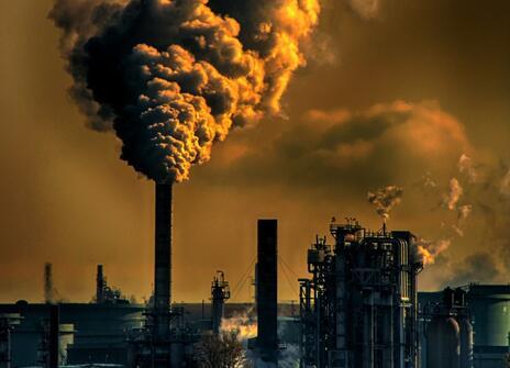 There is a landscape of chimney towers with lots of smoke coming out of them. The picture is a brown and yellow colour to show air pollution. 