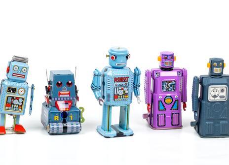 Five toy robots are stood in a line. They are all different shapes and colours - light blue, grey, purple and dark blue