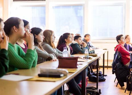 Children are sat at desks listening to a speaker