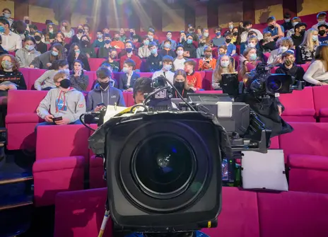 Image of a film camera lens with an audience sitting behind in a Theatre