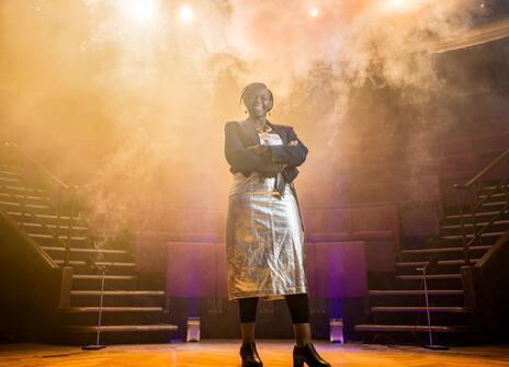 Maggie stands in the Theatre with dry ice and lights behind her