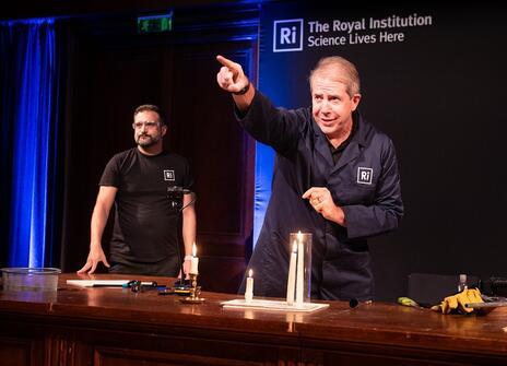 two men are at the faraday desk in the Ri theatre. a candle is alight on the desk.