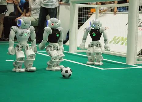 Three robots stand behind a football on an artificial football pitch, one of them is stood inside a goal.