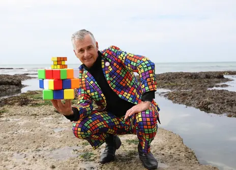 Andrew squatting down on a beach holding two rubik's cubes, one giant and one normal sized. He is wearing a rubik's cube patterned suit.
