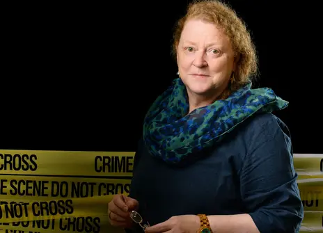 Sue Black, a white woman with curly hair, stands in front of yellow crime scene tape
