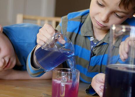 two boys are looking at glass containers filled with colourful liquid, one of the boys is pouring a blue liquid into a beaker of pink liquid