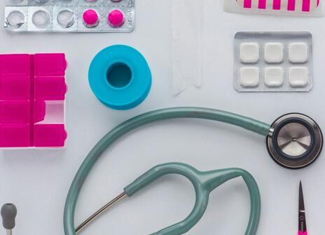 a stethoscope, pink pill box and other medical items are on a white table. 